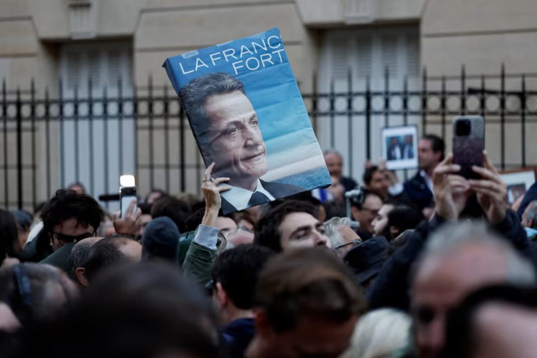 Pres 1 A supporter holds a poster with a portrait of Nicolas Sarkozy and the slogan "Strong France" as people attend a gathering called by the sons of former French President Nicolas Sarkozy in support of their father on the day he begins his five-year prison sentence at the Sante prison for criminal conspiracy over attempts to raise campaign funds from Libya, in Paris, France, October 21, 2025.